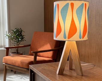 May include: A table lamp with a cylindrical shade featuring a repeating pattern of orange and light blue abstract shapes. The lamp has a light-colored wooden tripod base consisting of three angled legs that meet at the top to support the light fixture.