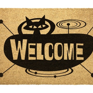 May include: A brown coir doormat with a black silhouette of a cat in a flying saucer with the word "WELCOME" printed on the saucer.