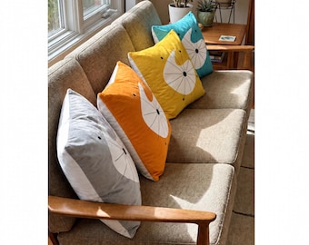 May include: Four square throw pillows arranged on a tan sofa. Each pillow features a solid color—gray, orange, yellow, and teal—with a white circular graphic and thin black radiating lines extending from a central point on the front surface.