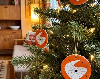 May include: Round ceramic ornaments hanging on a green pine tree. Each ornament features a bright orange background with a white crescent shape and thin black lines radiating from a central point, accented by small black dots at the ends of the lines.