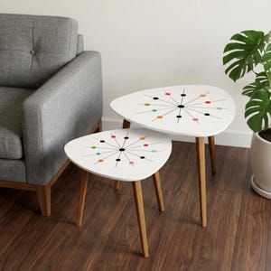 May include: A set of two white, kidney-shaped nesting tables with three light brown wooden legs each. The tabletops feature a black starburst pattern with colorful dots in red, orange, blue, green, and pink arranged in a radial design.