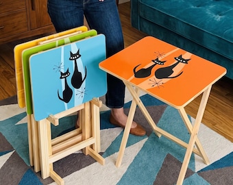 May include: A set of four rectangular folding snack tables with light wood frames. Three tables are stacked in a wooden stand, featuring yellow, green, and blue surfaces with black cat silhouettes and starburst designs. One orange table stands alone.