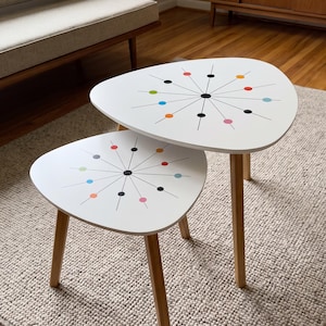 May include: A set of two white nesting tables with three light wood legs each. The tabletops feature a black starburst pattern with colorful dots in red, orange, yellow, green, blue, and pink arranged in a radial design on the white surface.