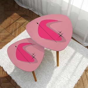 May include: Two pink, mid-century modern style side tables with a retro design. Each table has a triangular top with a curved fuchsia shape, black lines, and starburst accents. The tables have wooden legs and sit on a white shag rug.