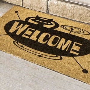 May include: A brown coir doormat with a black and white design of a flying saucer with the word "WELCOME" printed on it.