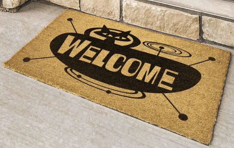 May include: A brown coir doormat with a black and white design of a flying saucer with the word "WELCOME" printed on it.