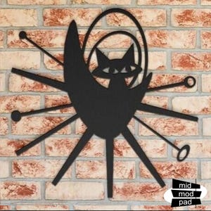May include: Black metal cat wall art in a mid-century modern style. The cat is set against a crescent moon shape, with radiating lines and circular accents. The art is mounted on a brick wall.