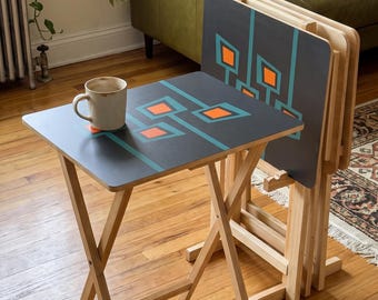 May include: A set of four folding snack tables with light wood legs and dark gray rectangular tops featuring a geometric pattern of teal lines and orange squares. A ceramic mug sits on the surface of the single unfolded table.