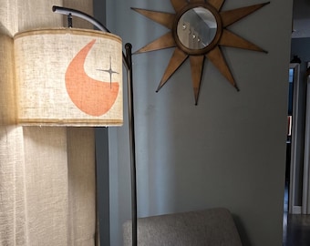 May include: A floor lamp with a cylindrical beige fabric shade featuring a burnt orange crescent moon and a black four-pointed star graphic. The lamp has a thin black metal stand. A gold-toned sunburst wall mirror hangs nearby.