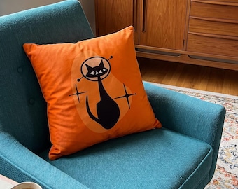 May include: A square orange throw pillow featuring a black graphic illustration of a cat wearing a space helmet, accented by two four-pointed star shapes and a light orange oval background shape printed on the front fabric surface.