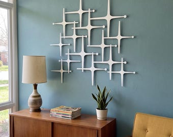 May include: A white wall sculpture featuring an interconnected grid of starburst shapes with rounded tips. The geometric design consists of multiple overlapping four-pointed elements arranged in a repeating pattern, mounted flat against a teal wall.