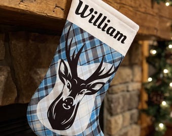 May include: A Christmas stocking featuring a blue and black plaid pattern with a white cuff displaying the name William in black text. A black and white graphic of a deer head with antlers is centered on the front of the fabric stocking.