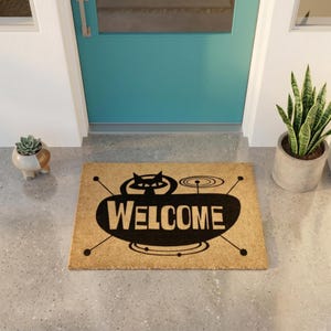 May include: A rectangular tan coir doormat featuring a black graphic design of a stylized cat head and the word WELCOME in bold capital letters, accented by thin black lines and circular shapes extending from the central black oval shape.
