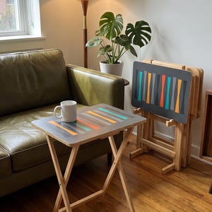 May include: A set of folding snack tables with light wood legs and rectangular tops featuring a gray surface decorated with a repeating pattern of vertical, tapered shapes in teal, orange, and gray. One table holds a white ceramic mug.