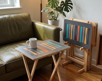 May include: A set of folding snack tables with light wood legs and rectangular tops featuring a gray surface decorated with a repeating pattern of vertical, tapered shapes in teal, orange, and gray. One table holds a white ceramic mug.