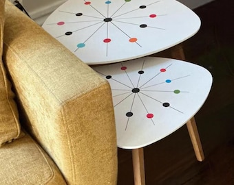 May include: Two white nesting tables with a rounded triangular shape and light wooden legs. The tabletops feature a black starburst pattern with colorful dots in red, blue, green, and black arranged along the radiating lines.