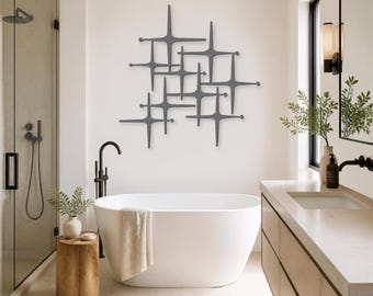 May include: A dark gray metal wall sculpture featuring a geometric starburst pattern with intersecting lines and rounded tips. The piece is mounted on a plain wall above a white freestanding bathtub in a bathroom setting with neutral decor.