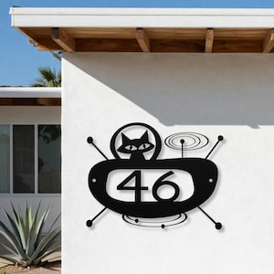 May include: A black metal house number sign featuring the number 46 centered within an oval frame. The design includes a stylized cat silhouette in a circular cutout above the number, accented by thin radiating lines and concentric circles.