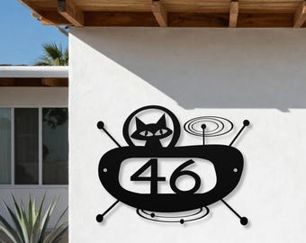 May include: A black metal house number sign featuring the number 46 centered within an oval frame. The design includes a stylized cat silhouette in a circular cutout above the number, accented by thin radiating lines and concentric circles.