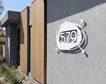May include: A white metal house number sign displaying the digits 579, featuring an abstract, rounded, television-like frame with four protruding antenna-style rods, mounted on a textured gray exterior wall of a modern building.