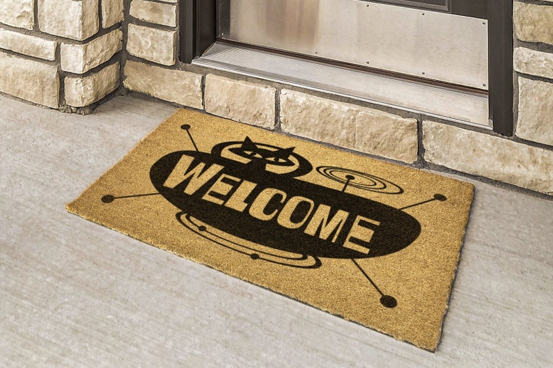 May include: A brown coir doormat with a black and white graphic of a flying saucer with the word "WELCOME" printed on it.