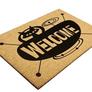 May include: A brown coir doormat with a black and white design of a retro spaceship with the word "WELCOME" printed on it.