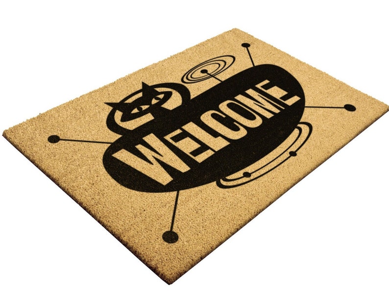 May include: A brown coir doormat with a black and white design of a retro spaceship with the word "WELCOME" printed on it.