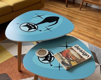 May include: Two light blue, kidney-shaped nesting tables with wooden legs. Each tabletop features a black graphic of a stylized cat with starburst accents. A small circular coaster and a book titled design rest on the lower table surface.