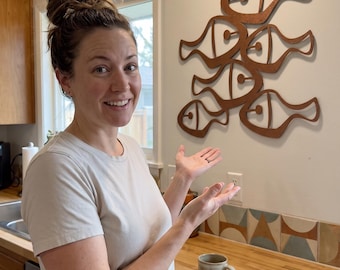 May include: A wall-mounted decorative art piece featuring a cluster of six stylized, interconnected fish shapes made from a brown, wood-toned material. The fish are arranged in a vertical formation with cut-out circular eye details on each fish.