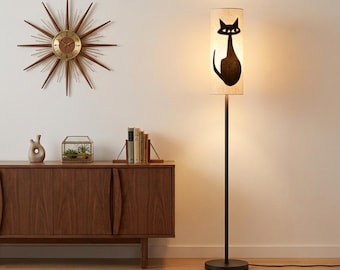 May include: A tall floor lamp with a cylindrical beige shade featuring a black cat silhouette graphic. The lamp has a thin black metal pole and a circular base. The light is illuminated, casting a warm glow from within the fabric shade.