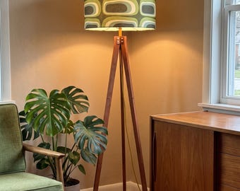 May include: A floor lamp with a wooden tripod base and a cylindrical fabric lampshade featuring a repeating geometric pattern in shades of olive green, light green, and dark gray. The lamp emits a warm glow from within the shade.