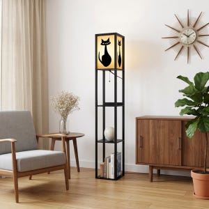 May include: A tall, black, rectangular floor lamp with integrated shelving. The top section features a light-colored shade with a black cat silhouette graphic on each side. The base includes three open shelves for storage, with books visible on the bottom shelf.