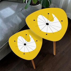 May include: A set of two yellow side tables with a rounded triangular shape. Each tabletop features a white abstract organic form overlaid with a black starburst pattern consisting of thin lines radiating from a central dot to small black circles.