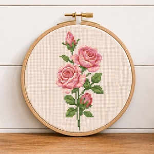 Könnte beinhalten: Ein runder Holzstickrahmen mit einem Kreuzstichmuster, das einen rosafarbenen Rosenzweig mit zwei voll erblühten Rosen und zwei Knospen zeigt. Das Motiv ist auf weißem Aida-Stoff gestickt und enthält grüne Blätter sowie Stängel.