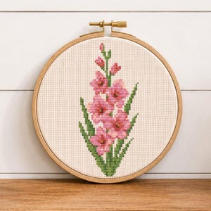 May include: A circular wooden embroidery hoop containing a cross-stitch pattern of a pink gladiolus flower with green leaves on a white fabric background. The embroidery hoop features a metal tightening screw at the top center of the frame.