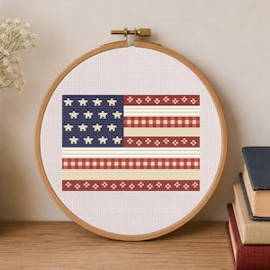 May include: A circular wooden embroidery hoop containing a cross-stitch pattern of the American flag. The design features a blue canton with white star-like shapes and horizontal stripes composed of red and white checkered and floral patterns.