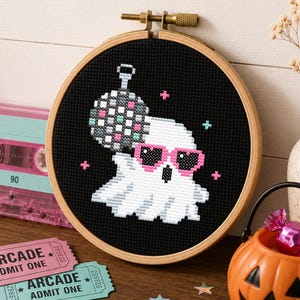 Può includere: Un telaio da ricamo rotondo in legno contenente un disegno a punto croce di un fantasma bianco che indossa occhiali da sole rosa a forma di cuore e tiene una palla da discoteca argentata su sfondo nero, con piccole stelle a punto croce.