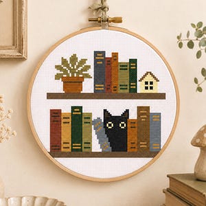 May include: A circular wooden embroidery hoop containing a cross-stitch pattern of two shelves. The top shelf features a potted plant, four books, and a small house. The bottom shelf displays five books and a black cat with two white eyes.