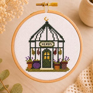 May include: A cross-stitch embroidery hoop with a greenhouse design. The design shows a green greenhouse with the word "HERBS" above the door, surrounded by potted plants in purple, green, and brown. The hoop is made of wood.