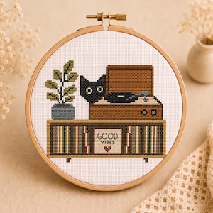 May include: A circular wooden embroidery hoop containing a cross-stitch pattern of a black cat sitting behind a brown record player on a cabinet. The cabinet features vertical stripes and a white square with the text GOOD VIBES and a small heart.