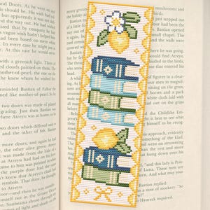 Può includere: Segnalibro ricamato con una pila di libri blu, verdi e bianchi, con dettagli di limoni e fiori. Il segnalibro è rettangolare con un bordo giallo e un fiocco in basso.