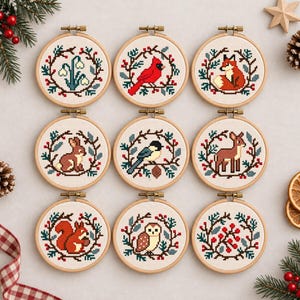 May include: Nine circular wooden embroidery hoops containing cross-stitch patterns of winter themes, including a cardinal, fox, rabbit, deer, squirrel, owl, chickadee, snowdrops, and red berries, each framed by a wreath of pine branches and holly.