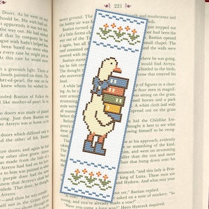 May include: Embroidered bookmark with a pixelated duck carrying books. The bookmark has a blue border with floral details. A charming accessory for book lovers, measuring approximately 20 cm in length.