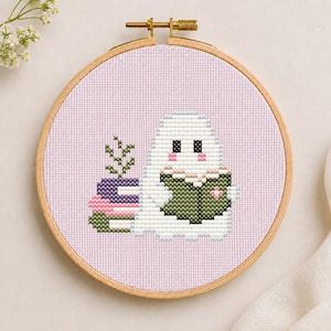 Può includere: Un ricamo a punto croce in un telaio rotondo di legno raffigurante un fantasma bianco con guance rosa che legge un libro verde scuro. Accanto al fantasma c'è una pila di tre libri viola, rosa e verdi, con una piccola pianta verde in cima.