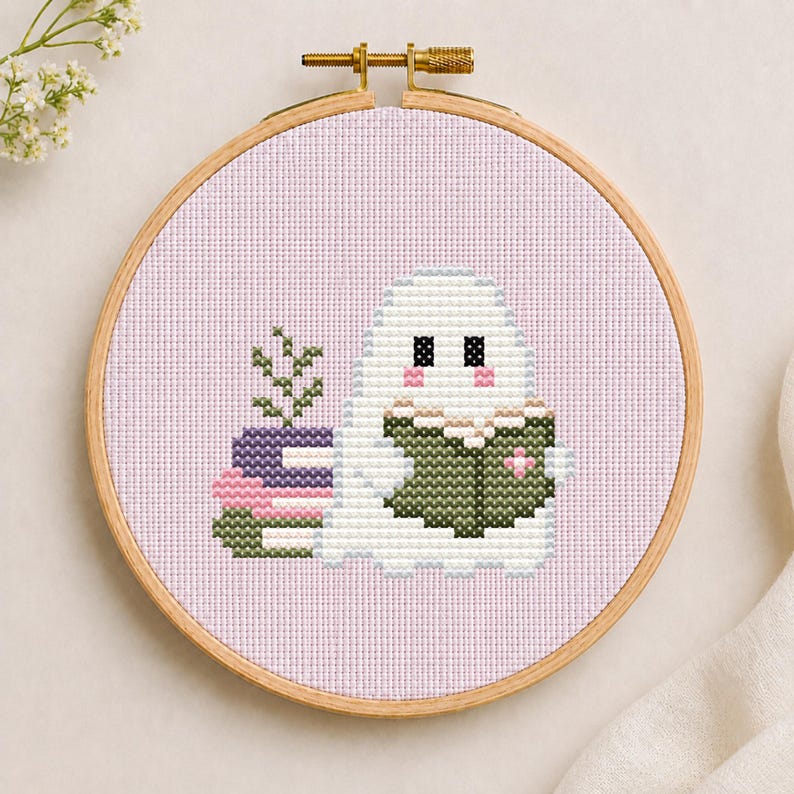 May include: A cross-stitch embroidery in a round wooden hoop featuring a white ghost with pink cheeks reading a dark green book. Beside the ghost is a stack of three books in purple, pink, and green, with a small green plant sprouting from the top.