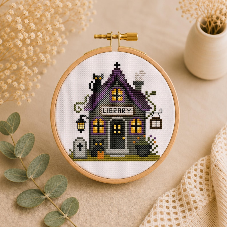 May include: A cross-stitch embroidery hoop featuring a spooky library scene. The house has a purple roof, glowing windows, and the word "LIBRARY" above the door. A black cat sits on the roof, and a jack-o'-lantern is in front.