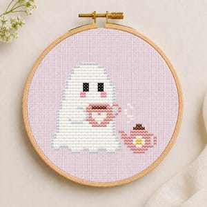 May include: A cross-stitch embroidery project in a round wooden hoop featuring a white ghost character holding a pink mug with a heart design. Next to the ghost is a small pink teapot with a white flower pattern on a light purple fabric background.