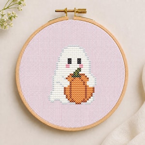 May include: A cross-stitch embroidery project in a round wooden hoop featuring a white ghost character with black eyes and pink cheeks holding an orange pumpkin with a green stem, all set against a light lavender fabric background.