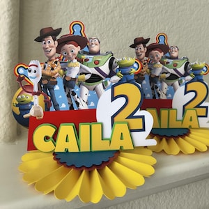 May include: A vibrant birthday cake topper with Toy Story characters. The design includes Woody, Jessie, Buzz Lightyear, and other characters, with the name "CAILA" and the number "2" displayed. The topper has a layered design with yellow, red, and blue accents.