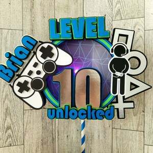 May include: A cake topper with a silver number 10, the words "LEVEL UNLOCKED", and a blue and green video game controller. The topper also features a blue and green gamer character with headphones and a blue and green "Brian" name tag.
